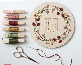 May include: A wooden embroidery hoop with a floral wreath design and the letter "H" embroidered in the center. The hoop is surrounded by embroidery floss in various colors, including pink, red, green, and gold.