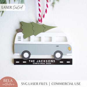 May include: A laser-cut Christmas ornament depicting a light blue and white camper van with a green Christmas tree. The ornament has black wheels and a black base with the text "THE JACKSONS CHRISTMAS 2024". It hangs from a red and white string.