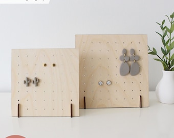 May include: Two wooden earring display stands with holes for hanging earrings. The stands are made of light wood and have a natural finish. The larger stand has a pair of grey clay earrings and a pair of white marble earrings. The smaller stand has a pair of gold earrings and a pair of cactus-shaped earrings.