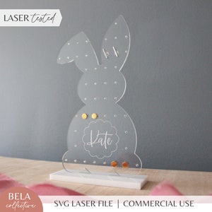 May include: A clear acrylic bunny-shaped earring holder with a white base. The holder has a circle with the name "Kate" inscribed in it. There are several gold-colored earrings on the holder.