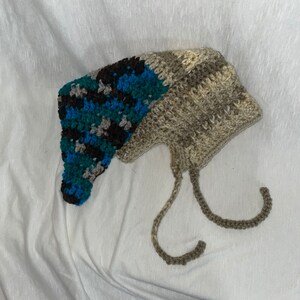 May include: A handmade crochet hat with a unique design. The hat features a section of teal, blue, black, and gray yarn, and a section of tan and brown yarn. The hat has two long ties. The hat is likely for babies or children.