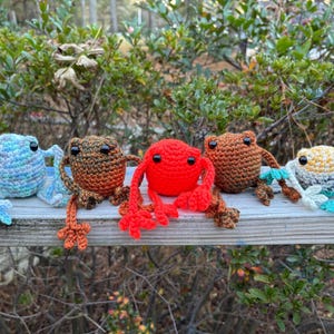 May include: Five crocheted frog figurines in various colors, including blue, brown, and red, are arranged on a wooden surface. Each frog has black eyes and detailed legs. The frogs are handmade and appear to be a craft item.