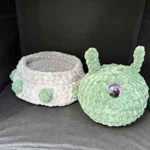 May include: A crocheted gray and green stuffed animal with a single purple eye. The animal is shaped like a slug or caterpillar and has a small, gray, crocheted bowl attached to its back.