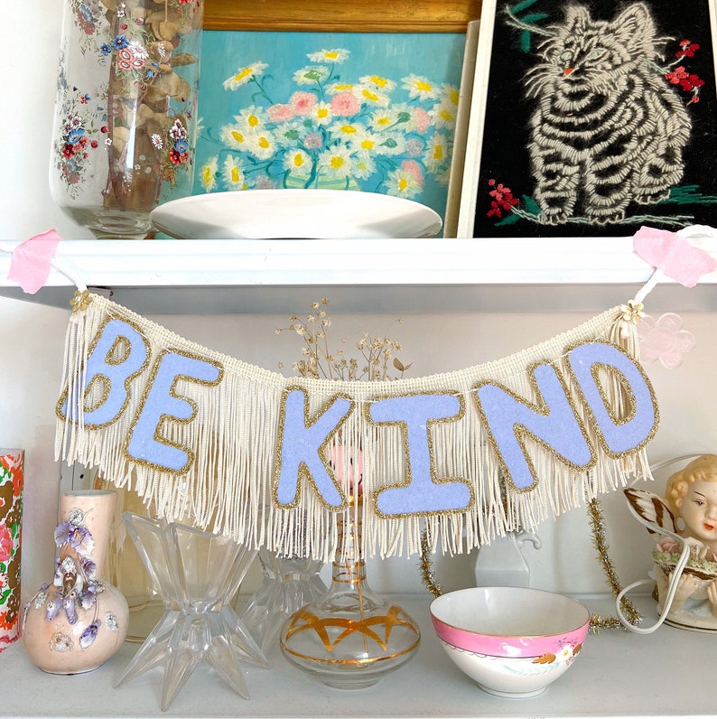 Puede incluir: Una pancarta blanca con ribete de flecos deletrea "BE KIND" en letras de fieltro lavanda. La pancarta est&aacute; colgada en un estante blanco con otros art&iacute;culos decorativos.