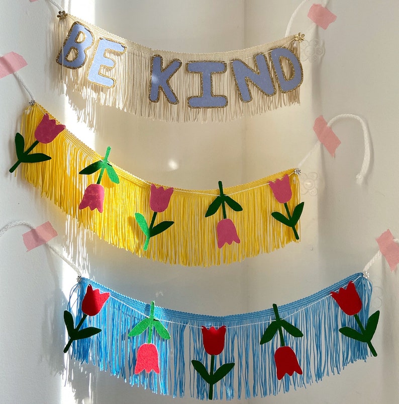 Puede incluir: Un banner con las palabras "Be Kind" en letras azul claro con brillo sobre un fondo blanco con flecos. El banner est&aacute; colgado de una pared blanca. Debajo del banner hay dos banners m&aacute;s con flecos amarillos y azules y formas de flores rojas y rosas.
