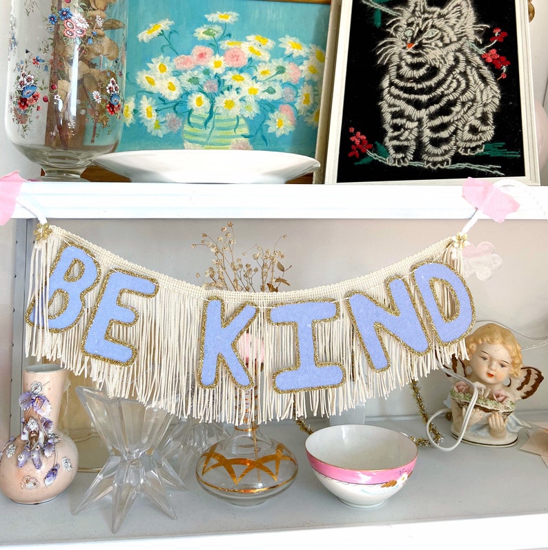 Puede incluir: Una pancarta blanca con flecos con las palabras "Be Kind" en letras azul claro. La pancarta est&aacute; colgada en un estante blanco con otros art&iacute;culos decorativos.