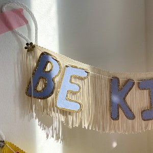 Puede incluir: Una pancarta de flecos blancos con las palabras "Be Kind" en letras lavanda con detalles de purpurina dorada.