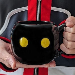 May include: Black ceramic mug with a round shape and two yellow eyes. The mug has a handle on the right side.