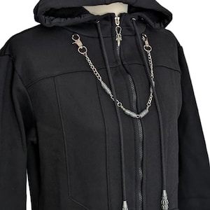K&ouml;nnte beinhalten: Ein schwarzer Kapuzenpullover mit Rei&szlig;verschluss, Kapuze mit Kordelzug, durchgehendem Frontrei&szlig;verschluss und einer dekorativen silberfarbenen Metallkettenverzierung am Kragenbereich, die kleine zylindrische Perlen und Karabinerverschl&uuml;sse aufweist.