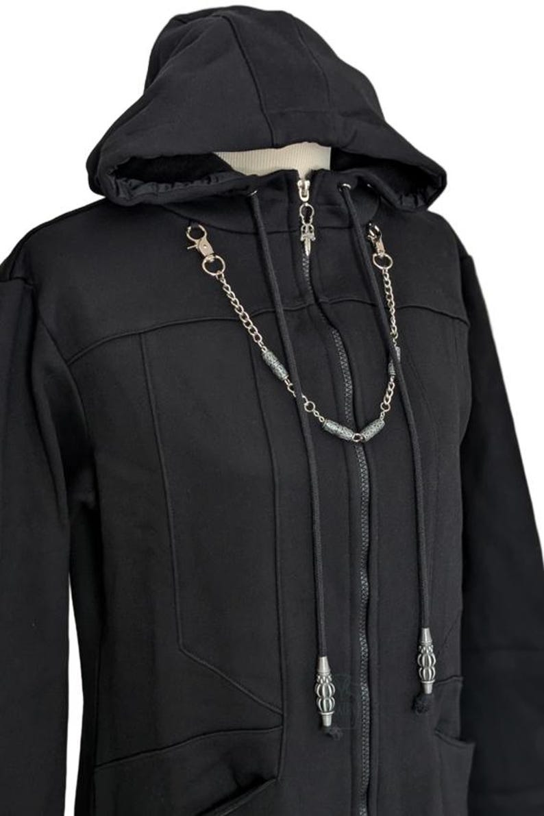 K&ouml;nnte beinhalten: Ein schwarzer Kapuzenpullover mit Rei&szlig;verschluss, Kapuze mit Kordelzug, durchgehendem Frontrei&szlig;verschluss und einer dekorativen silberfarbenen Metallkettenverzierung am Kragenbereich, die kleine zylindrische Perlen und Karabinerverschl&uuml;sse aufweist.