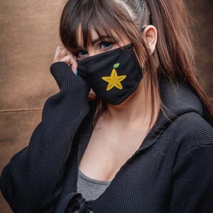 May include: A woman with long dark hair wearing a black hooded sweater and a black face mask with a yellow starfruit design.