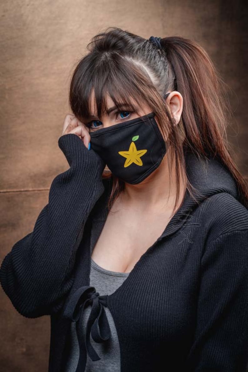 May include: A woman with long dark hair wearing a black hooded sweater and a black face mask with a yellow starfruit design.