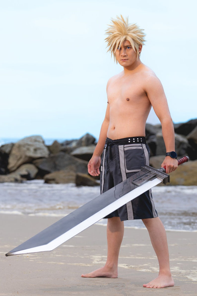 May include: A man wearing black swim trunks and a blonde wig is holding a large, black sword. He is standing on a sandy beach with rocks in the background.