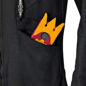 K&ouml;nnte beinhalten: Ein schwarzer Kapuzenpullover mit Rei&szlig;verschluss und einer Vordertasche, in der sich ein rot-gelbes kronenf&ouml;rmiges Objekt befindet. Der Pullover hat einen Kordelzug mit silbernem Metallabschluss und ein kleines gepr&auml;gtes Logo am Saum.