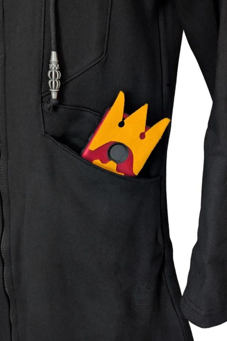 K&ouml;nnte beinhalten: Ein schwarzer Kapuzenpullover mit Rei&szlig;verschluss und einer Vordertasche, in der sich ein rot-gelbes kronenf&ouml;rmiges Objekt befindet. Der Pullover hat einen Kordelzug mit silbernem Metallabschluss und ein kleines gepr&auml;gtes Logo am Saum.