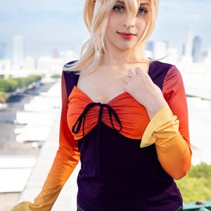 May include: A long-sleeved top with a color gradient from orange to yellow, paired with a deep purple bodice. The top features a gathered orange section at the chest, tied with a black string. The outfit is completed with black pants and a belt.