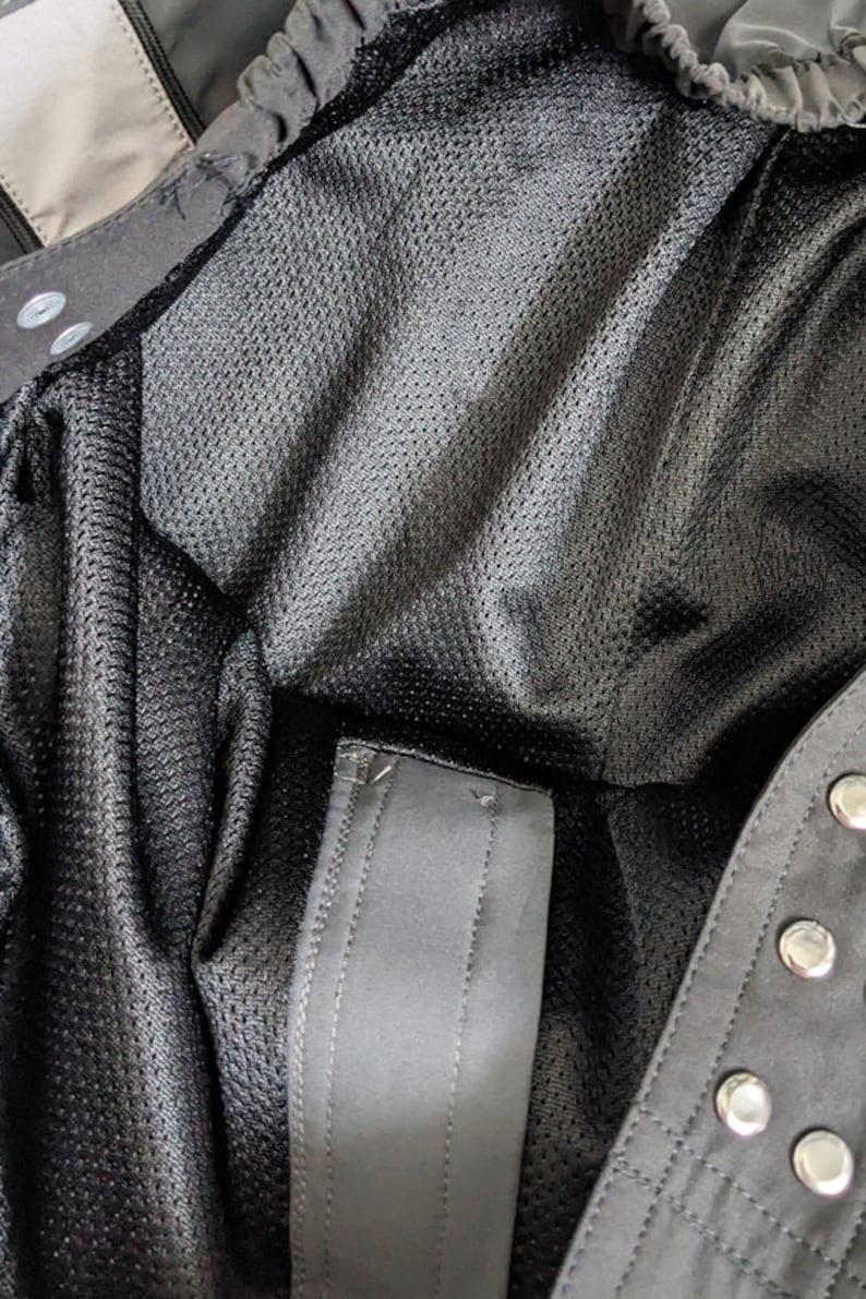 May include: Close-up of the inside of a black jacket with a mesh lining and a zipper closure. The jacket has a black strap with a snap closure.