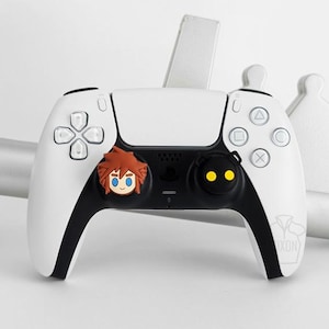 May include: White and black PlayStation 5 controller with a cartoon character face on the left side and a black button with yellow dots on the right. The controller has a logo on the bottom right.