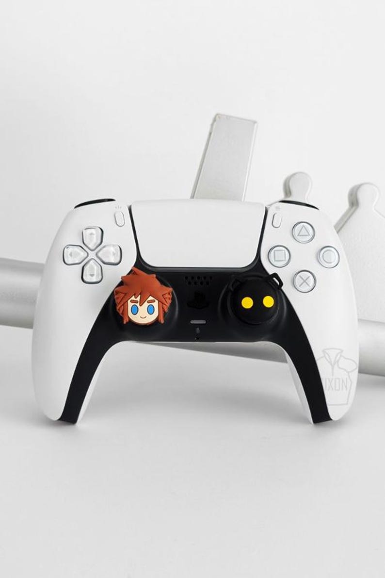 May include: White and black PlayStation 5 controller with a cartoon character face on the left side and a black button with yellow dots on the right. The controller has a logo on the bottom right.