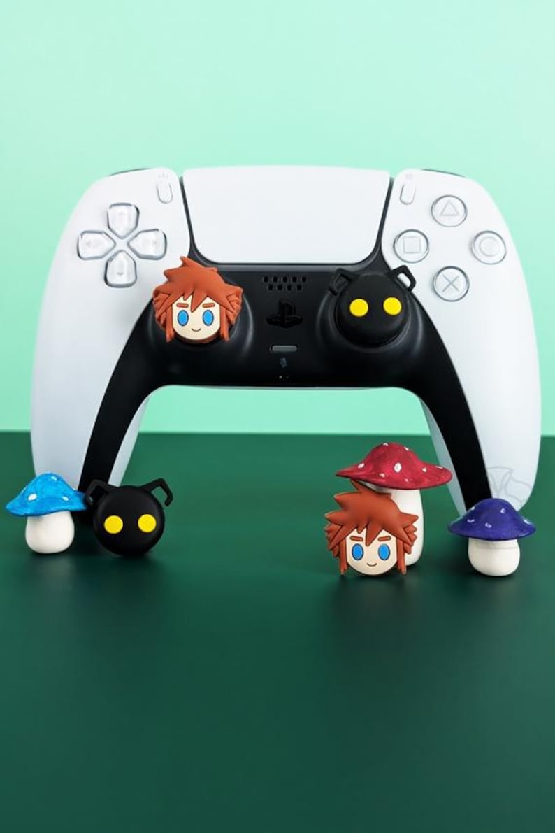 May include: A white and black game controller with character-themed accessories. The accessories include mushroom and character designs in various colors. The controller is on a green surface.