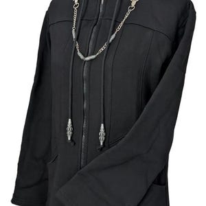 K&ouml;nnte beinhalten: Ein schwarzer Kapuzenpullover mit Rei&szlig;verschluss, durchgehendem Frontrei&szlig;verschluss, Kapuze mit langen Kordelz&uuml;gen und einer dekorativen silberfarbenen Metallkette am Ausschnitt. Das Kleidungsst&uuml;ck hat lange &Auml;rmel und seitliche Taschen.
