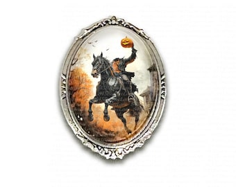 May include: Oval-shaped silver-toned brooch featuring a Halloween-themed illustration. The image depicts a headless figure on a black horse holding a glowing orange pumpkin. The background shows a spooky landscape with trees and a building.
