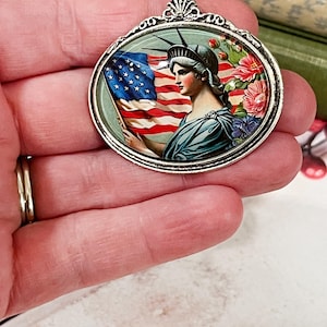 May include: Oval pendant with a silver-tone frame. The pendant features a patriotic design with the Statue of Liberty holding an American flag, surrounded by flowers. The pendant is a vintage-style accessory.