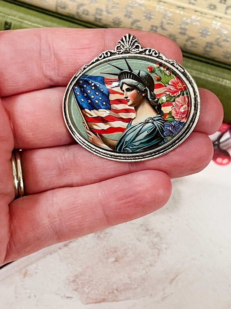 May include: Oval pendant with a silver-tone frame. The pendant features a patriotic design with the Statue of Liberty holding an American flag, surrounded by flowers. The pendant is a vintage-style accessory.