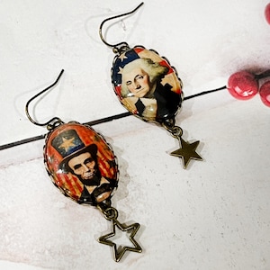 Puede incluir: Pendientes patri&oacute;ticos con colgantes ovalados que representan a Abraham Lincoln y George Washington. Los colgantes est&aacute;n enmarcados en metal color bronce y cuelgan con dijes en forma de estrella. El fondo incluye colores rojo, blanco y azul.