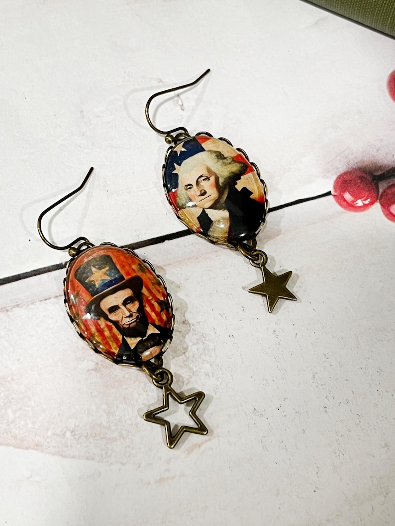 Puede incluir: Pendientes patri&oacute;ticos con colgantes ovalados que representan a Abraham Lincoln y George Washington. Los colgantes est&aacute;n enmarcados en metal color bronce y cuelgan con dijes en forma de estrella. El fondo incluye colores rojo, blanco y azul.