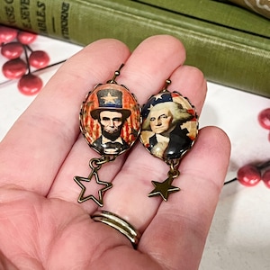 Puede incluir: Pendientes ovalados con retratos de Abraham Lincoln y George Washington, con colgantes de estrella. Los pendientes tienen una est&eacute;tica vintage, con metal color bronce e im&aacute;genes patri&oacute;ticas. El fondo incluye libros y bayas rojas.
