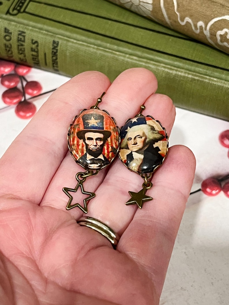 Puede incluir: Pendientes ovalados con retratos de Abraham Lincoln y George Washington, con colgantes de estrella. Los pendientes tienen una est&eacute;tica vintage, con metal color bronce e im&aacute;genes patri&oacute;ticas. El fondo incluye libros y bayas rojas.