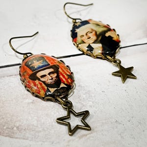 Puede incluir: Un par de pendientes de estilo antiguo. Cada pendiente tiene un colgante ovalado con im&aacute;genes de figuras hist&oacute;ricas, incluyendo a Abraham Lincoln y George Washington. Cada pendiente tiene un dije de estrella de color bronce.