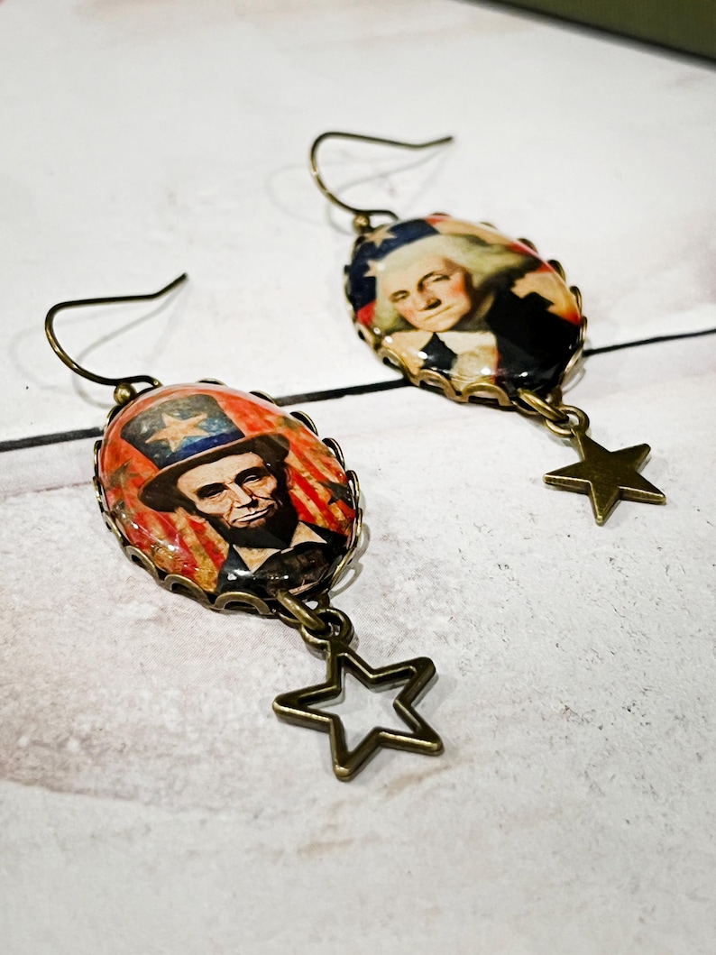 Puede incluir: Un par de pendientes de estilo antiguo. Cada pendiente tiene un colgante ovalado con im&aacute;genes de figuras hist&oacute;ricas, incluyendo a Abraham Lincoln y George Washington. Cada pendiente tiene un dije de estrella de color bronce.