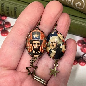 Puede incluir: Un par de pendientes patri&oacute;ticos con colgantes ovalados con im&aacute;genes de Abraham Lincoln y George Washington. Los colgantes est&aacute;n enmarcados en un metal color bronce y cuelgan con dijes de estrella. El fondo incluye elementos rojos, blancos y azules.