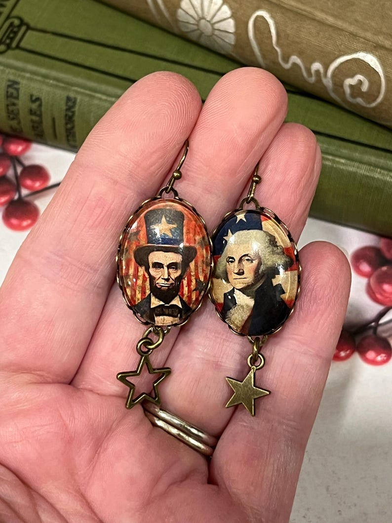 Puede incluir: Un par de pendientes patri&oacute;ticos con colgantes ovalados con im&aacute;genes de Abraham Lincoln y George Washington. Los colgantes est&aacute;n enmarcados en un metal color bronce y cuelgan con dijes de estrella. El fondo incluye elementos rojos, blancos y azules.