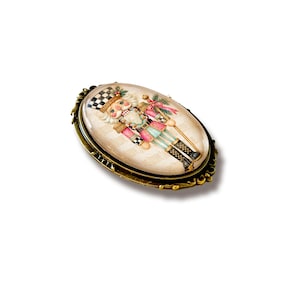 May include: An oval brooch featuring a nutcracker illustration with a black and white checkered hat, pink jacket, and light blue trousers. The image is set under a clear glass dome and framed in an ornate, antique-style bronze metal setting.