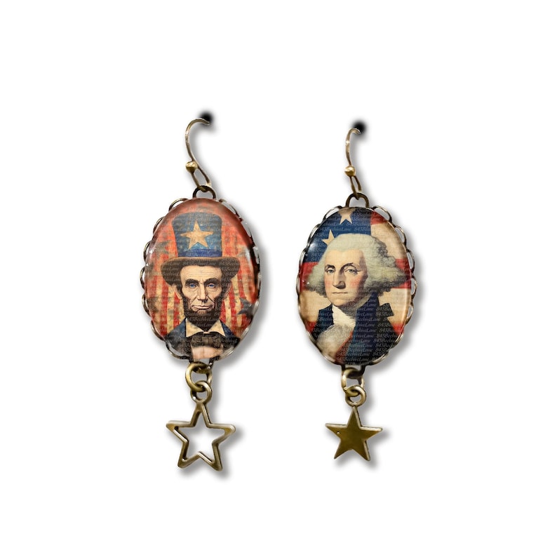 Puede incluir: Un par de pendientes patri&oacute;ticos con colgantes ovalados que representan a Abraham Lincoln y George Washington sobre un fondo de la bandera estadounidense. Cada pendiente tiene un dije de estrella.