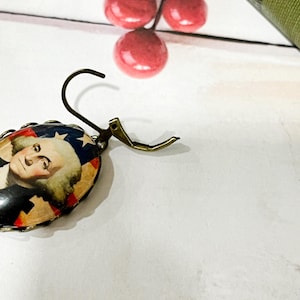 Puede incluir: Pendiente ovalado con un dise&ntilde;o patri&oacute;tico que presenta un retrato de George Washington, sobre un fondo rojo, blanco y azul. El pendiente tiene un gancho y cierre de color bronce.