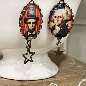 Puede incluir: Pendientes patri&oacute;ticos con colgantes ovalados que representan a Abraham Lincoln y George Washington. Los colgantes est&aacute;n enmarcados con un borde decorativo y tienen colgantes de estrella. Dise&ntilde;o vintage.