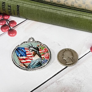May include: An oval pendant featuring the Statue of Liberty holding an American flag, surrounded by floral accents. A silver quarter coin is next to the pendant. The pendant is set against a white background with books.