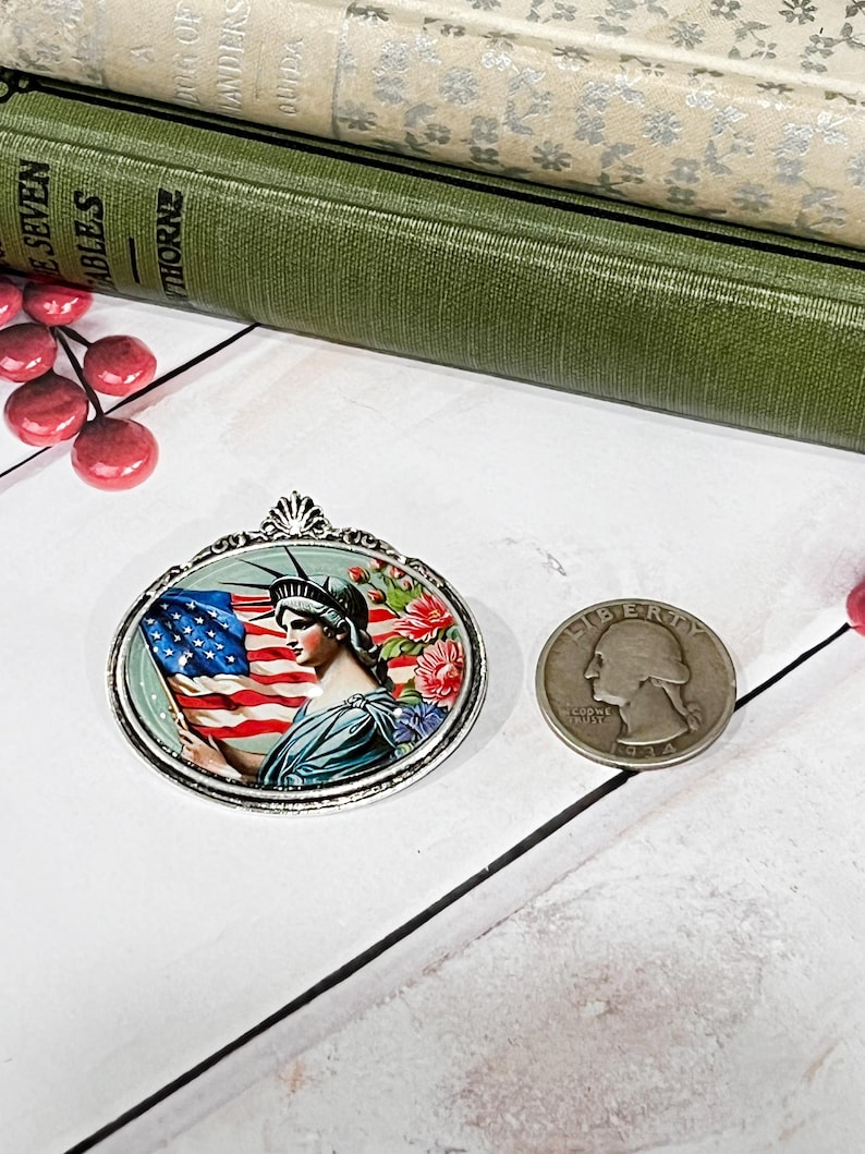 May include: An oval pendant featuring the Statue of Liberty holding an American flag, surrounded by floral accents. A silver quarter coin is next to the pendant. The pendant is set against a white background with books.