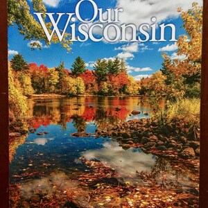 May include: A magazine cover titled "Our Wisconsin" features a scenic autumn landscape. The image showcases a lake reflecting colorful trees and a bright blue sky with clouds. The cover also includes the publication date.