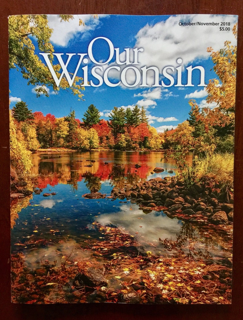 May include: A magazine cover titled "Our Wisconsin" features a scenic autumn landscape. The image showcases a lake reflecting colorful trees and a bright blue sky with clouds. The cover also includes the publication date.