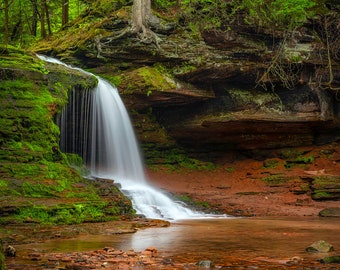 Puede incluir: Una cascada cae sobre rocas cubiertas de musgo en un exuberante bosque verde. El agua es clara y las rocas son de color marrón rojizo.