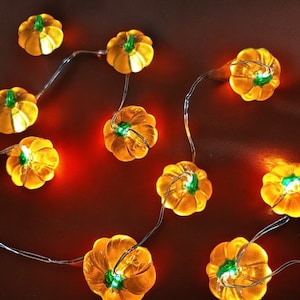 May include: A string of decorative lights featuring small, orange pumpkin-shaped bulbs with green stems. The lights are strung on a thin, silver wire, and are arranged on a dark orange surface. The pumpkins are lit, creating a warm, festive glow.