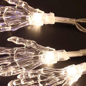 May include: A string of decorative lights featuring multiple clear plastic skeleton hand shapes. Each hand is connected to a thin wire and contains a small, glowing white LED bulb at the wrist, creating a series of illuminated skeletal forms.