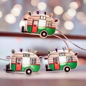 May include: A string of fairy lights with three white camper van shaped lights. Each light has a green and white exterior with a red door and windows.