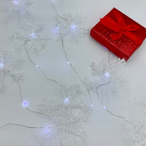 May include: A red gift box with a matching ribbon sits next to a string of snowflake lights. The lights are white and the snowflakes are clear. The background is white.