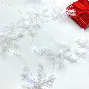 May include: A string of white snowflake lights and a small red gift box with a matching ribbon. The lights are strung on a thin silver wire and are scattered across a white surface, creating a festive holiday scene.